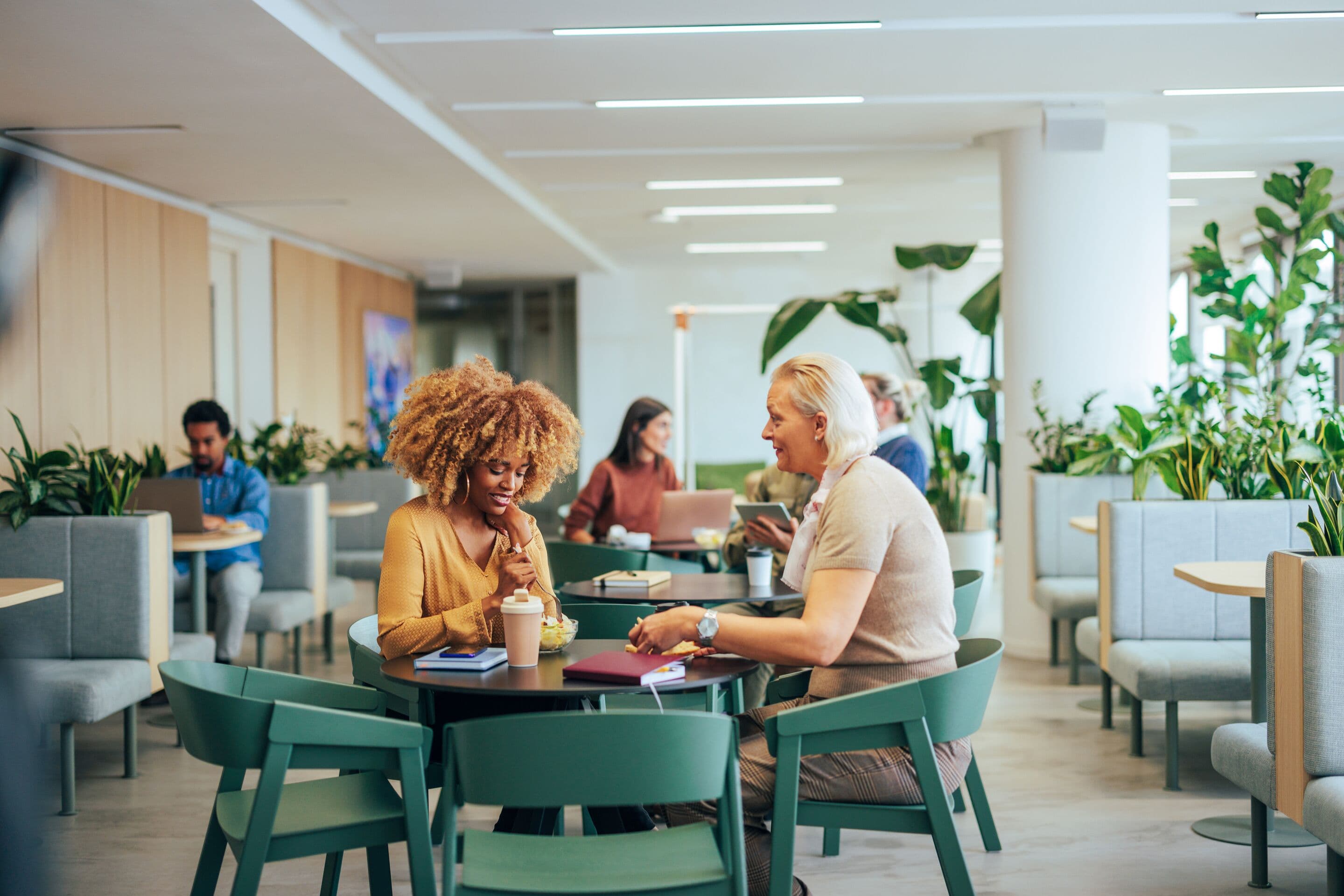 Two female coworkers are enjoying their lunch break at work, sitting at the table in the cafeteria and talking to each other.