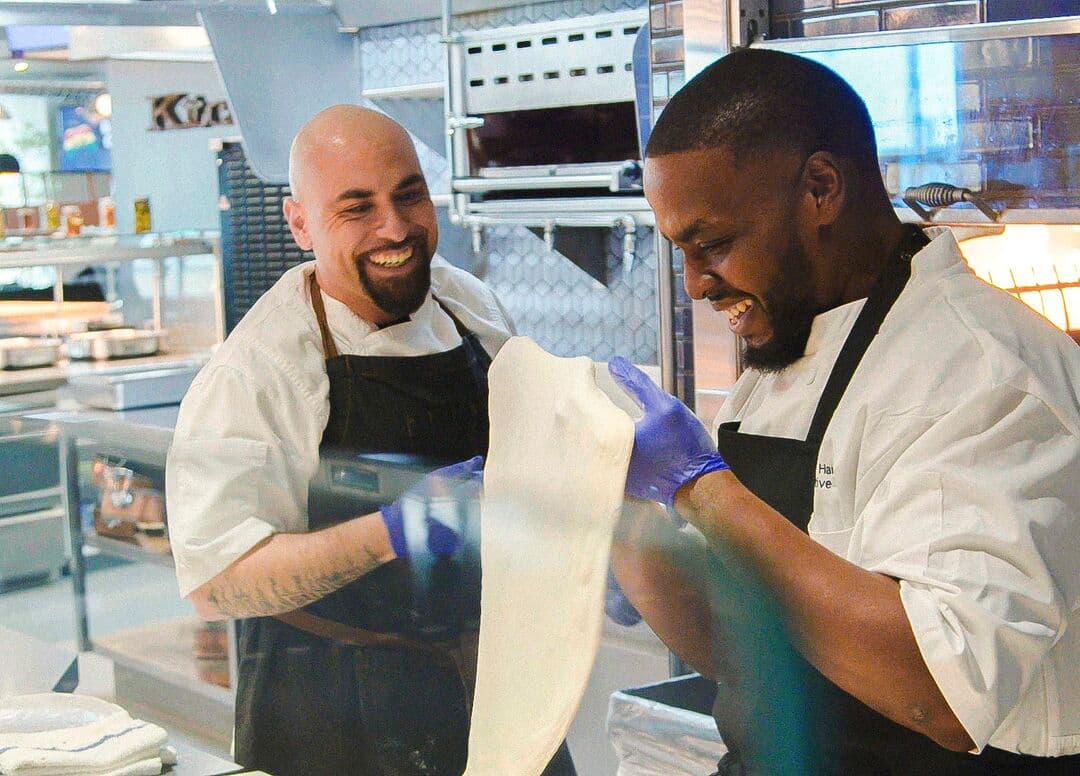 A professional chef guides a colleague in crafting artisan pizza dough that they will cook in a wood-fired pizza oven.