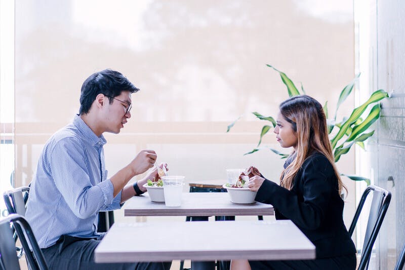 A pair of diverse colleagues sit and have a healthy salad lunch together. They are professionally dressed and chatting as they dig into their food. One is a Malay Asian woman, the other a Korean man.
